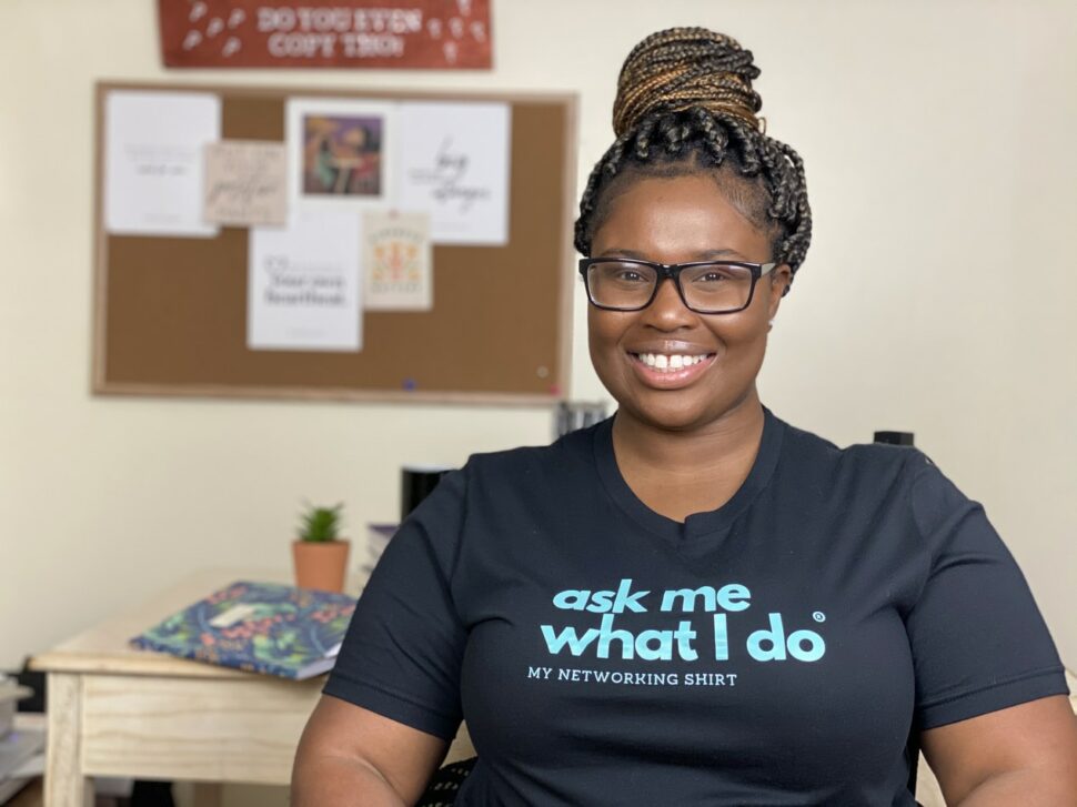 A Black woman smiles at the camera with a shirt that says "Ask me what I do: My networking shirt." 